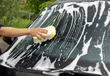 Hand using a yellow sponge to wash a soapy car windshield during car detailing.