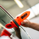 A hand applying Glaco glass water repellent using an orange applicator on a car window.