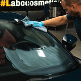 A person using a microfiber cloth to apply Labocosmetica glass polish to a car windshield.