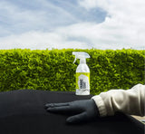 Fabric Coat spray bottle for car interior protection, shown with gloved hand on black upholstery against green hedge